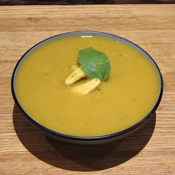 A bowl of yellow Indian soup, garnished with paneer and a mint leaf, on a wooden table.