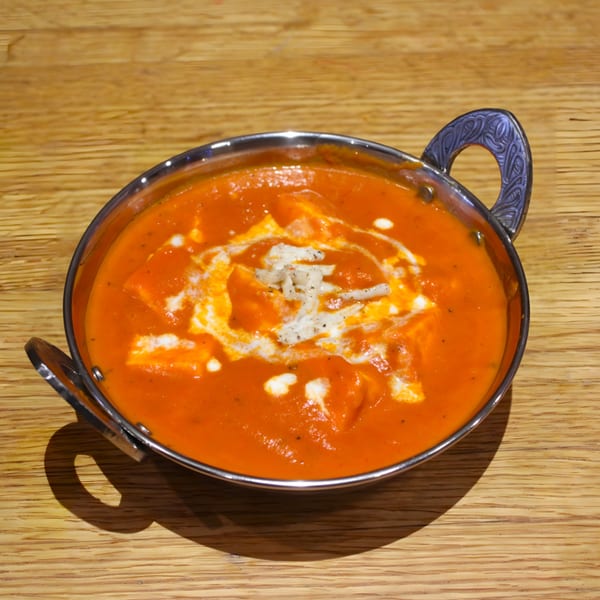 A vibrant orange Indian curry, possibly paneer butter masala, in a silver serving bowl on a wooden table.