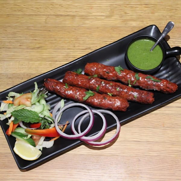 Four Mint Masala Indian seekh kebabs with green chutney, salad, and red onion on a black plate.